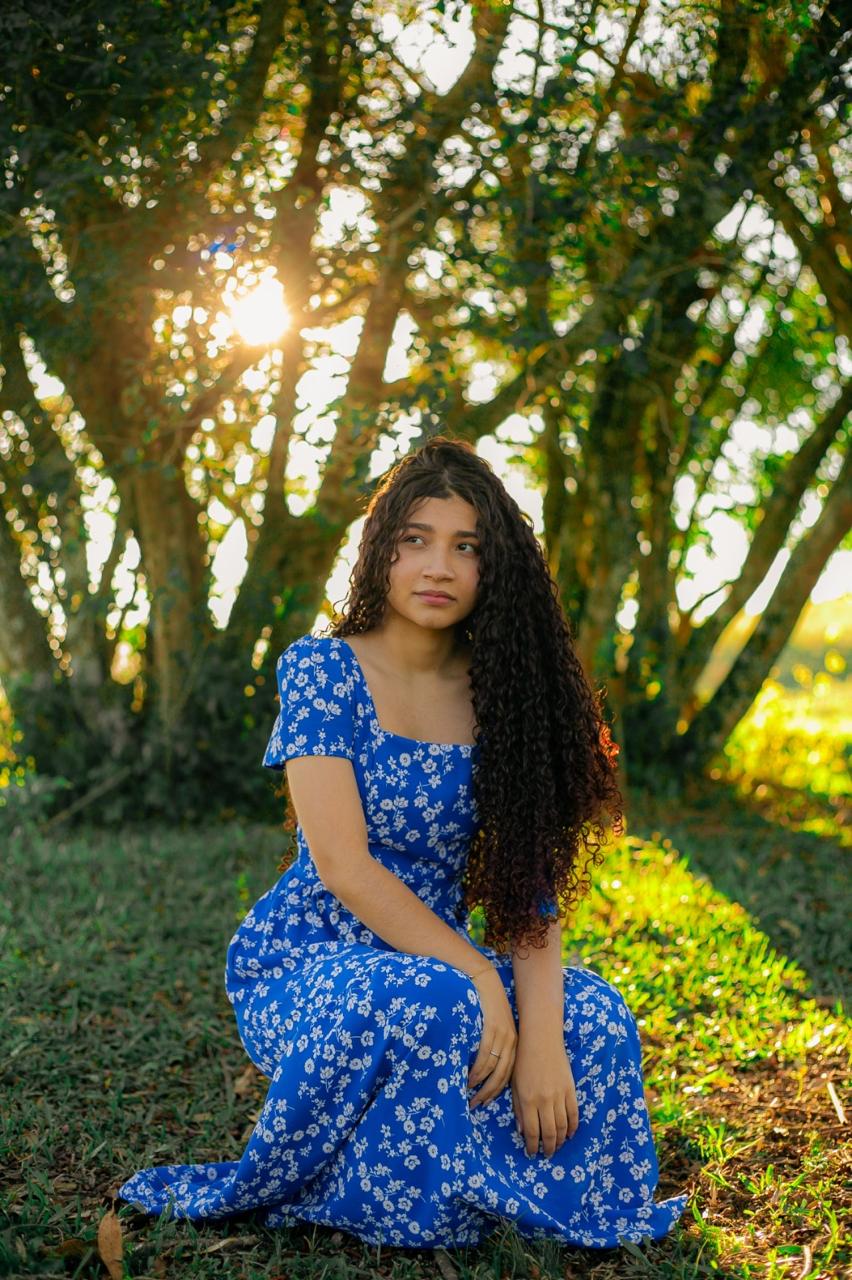 Jovem de vestido azul florido sentada entre árvores ao pôr do sol