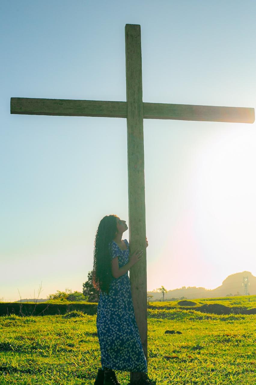 Jovem de vestido azul junto a uma cruz de madeira ao ar livre